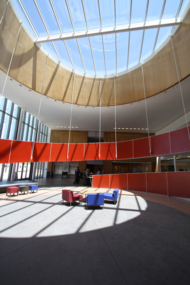 A large room with a large glass ceiling, a group of chairs in a wide open space, and a suspended red helical ramp winding up to second floor.
