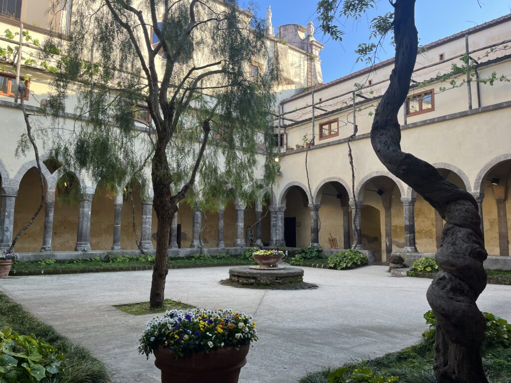 A courtyard is surrounded with the arched walkway of a tall building made of tan stone. Flowers, trees, and a small fountain fill the courtyard.