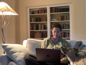 Alison Cook-Sather, seated on a couch with laptop in lap and her dog sitting next to her