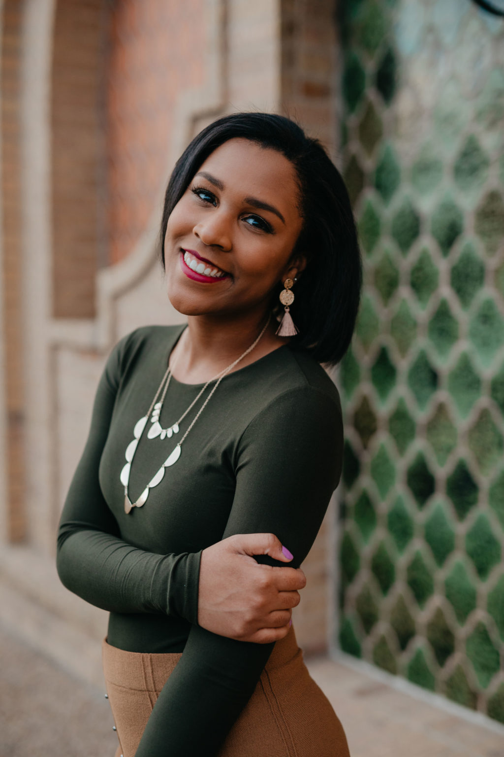 A smiling woman stands looking at the camera with one arm across her waist, grasping her other arm.