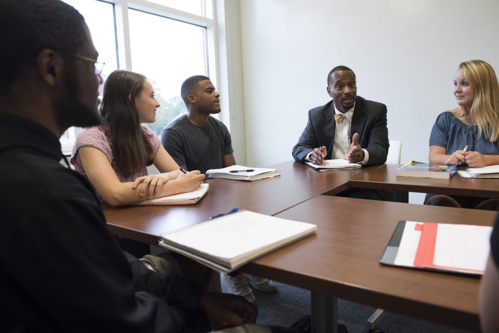 A professor and group of students talk together around a small conference table.