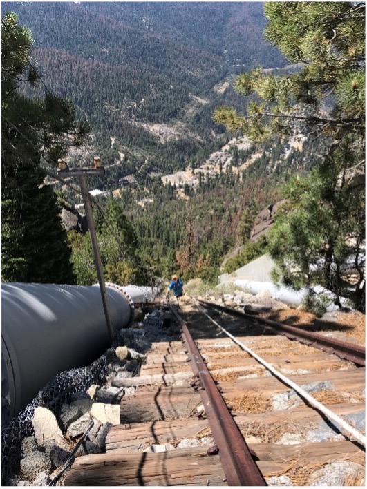 Boy in orange hat and clue shirt climbing up a train track with penstock pipes on either side. It is very steep and the town of Big Creek, CA is in the background.