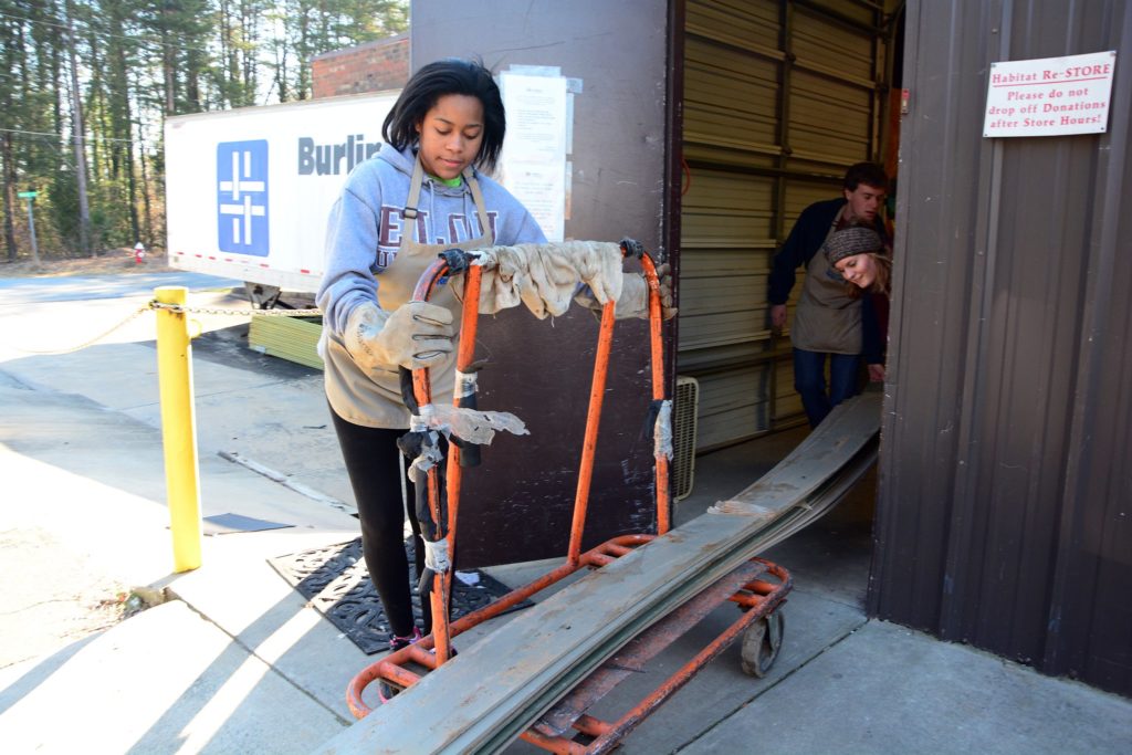 A student in an apron and heavy gloves works at the Habitat Re-Store, moving building materials on a cart.