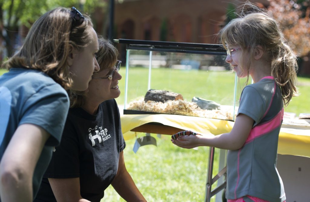 Two women squat next to a young child who holds a snack in her hands. The snake's tank is visible on a table behind them.