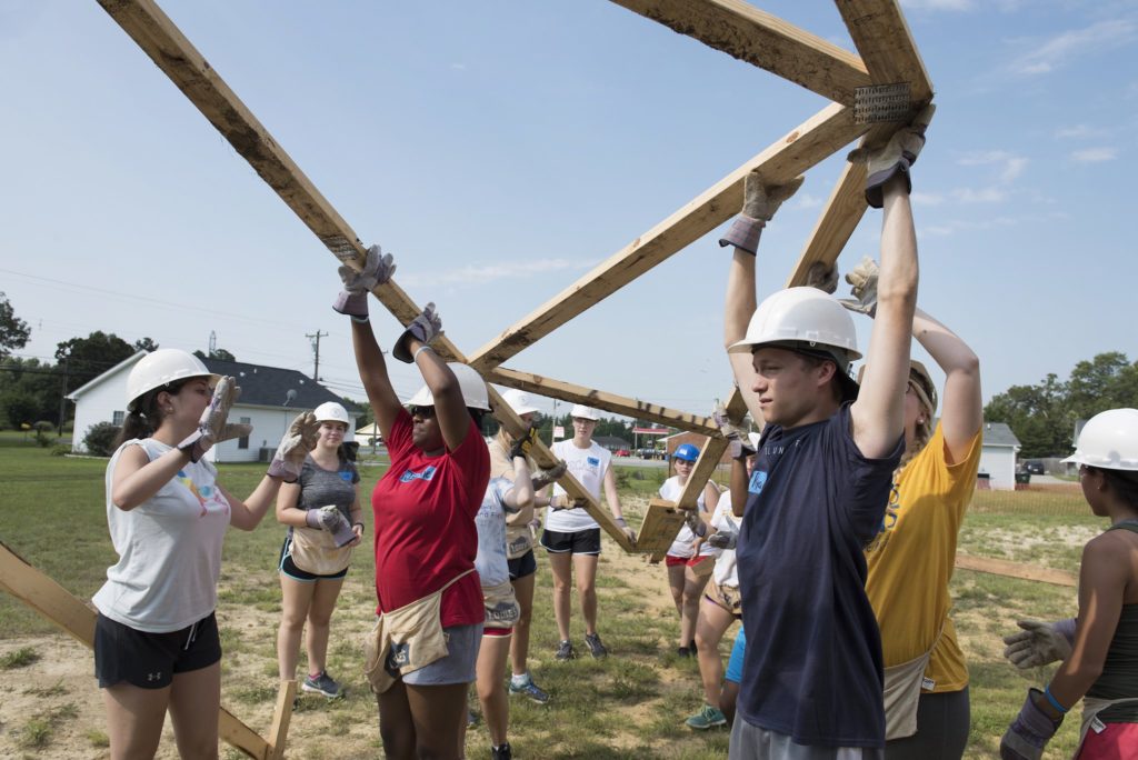 Students in hard hats work on a construction site, raising a wooden frame.