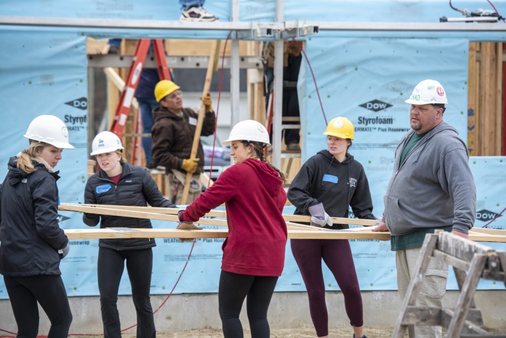 Several students in hard hats work on a construction site, carrying the wooden frame of a wall. 