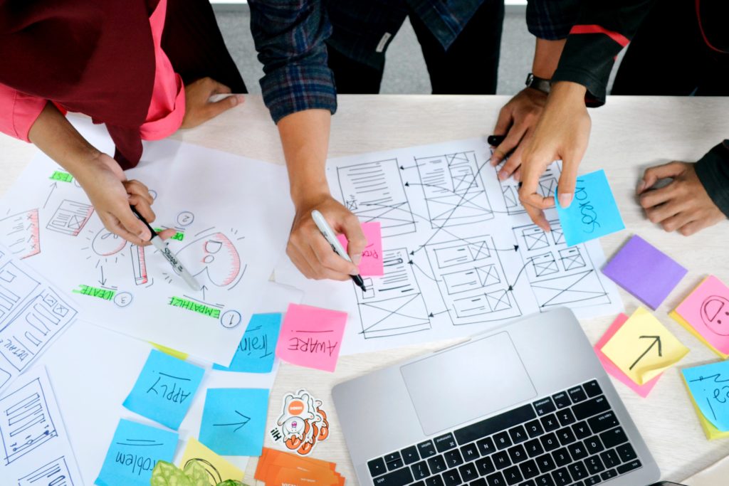 A desk covered with hand-drawn wireframes, sticky notes, and a computer. The hands of three people are visible working on the papers.