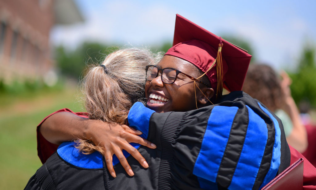 A student in graduation robes hugs a professor.