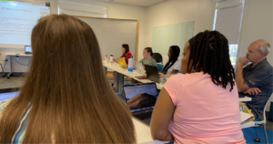 Six people are gathered at tables with laptops open in front of them. They are looking at a projection screen in the top left corner.