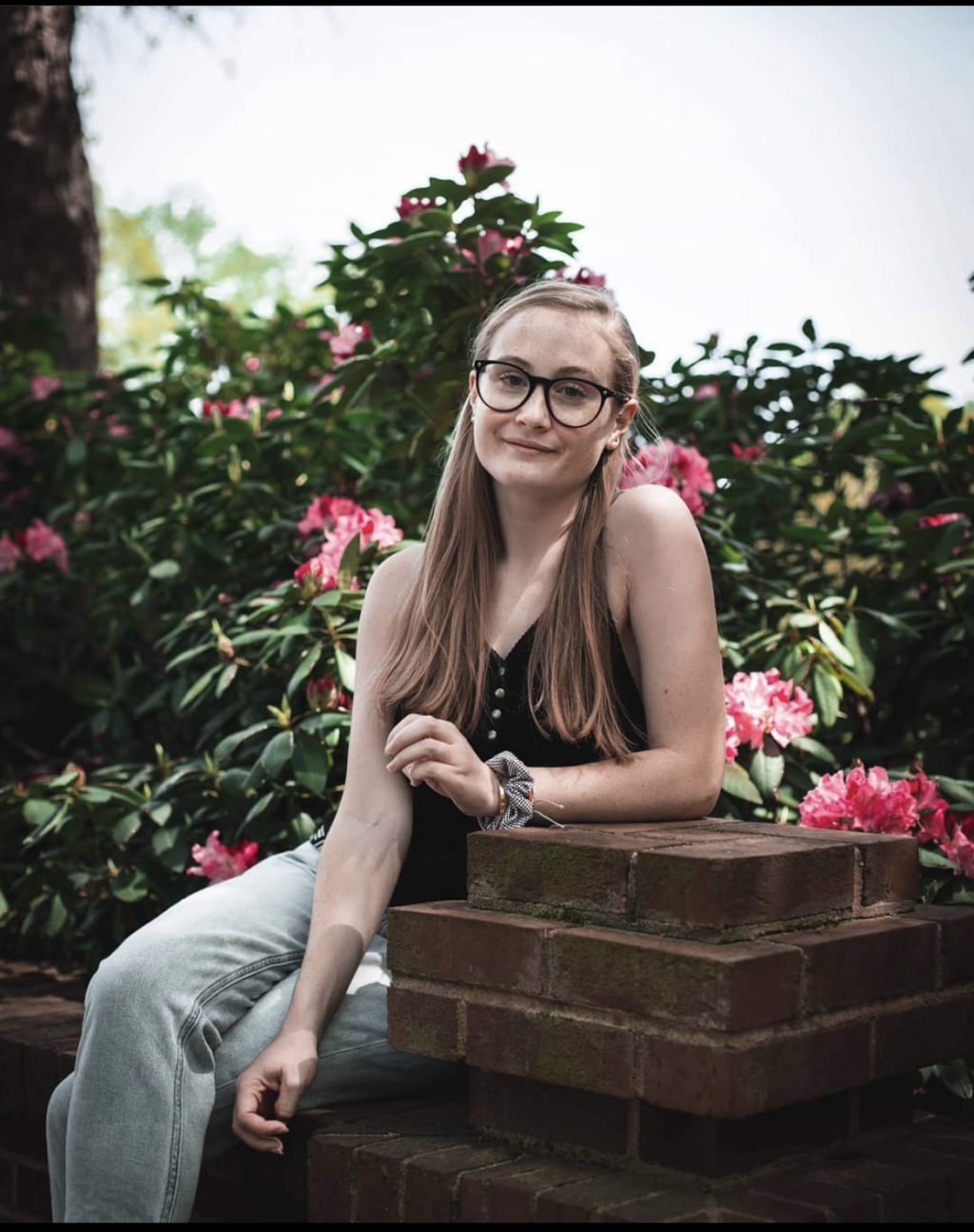 A woman sits on a low brick wall. In the background are flowering bushes.