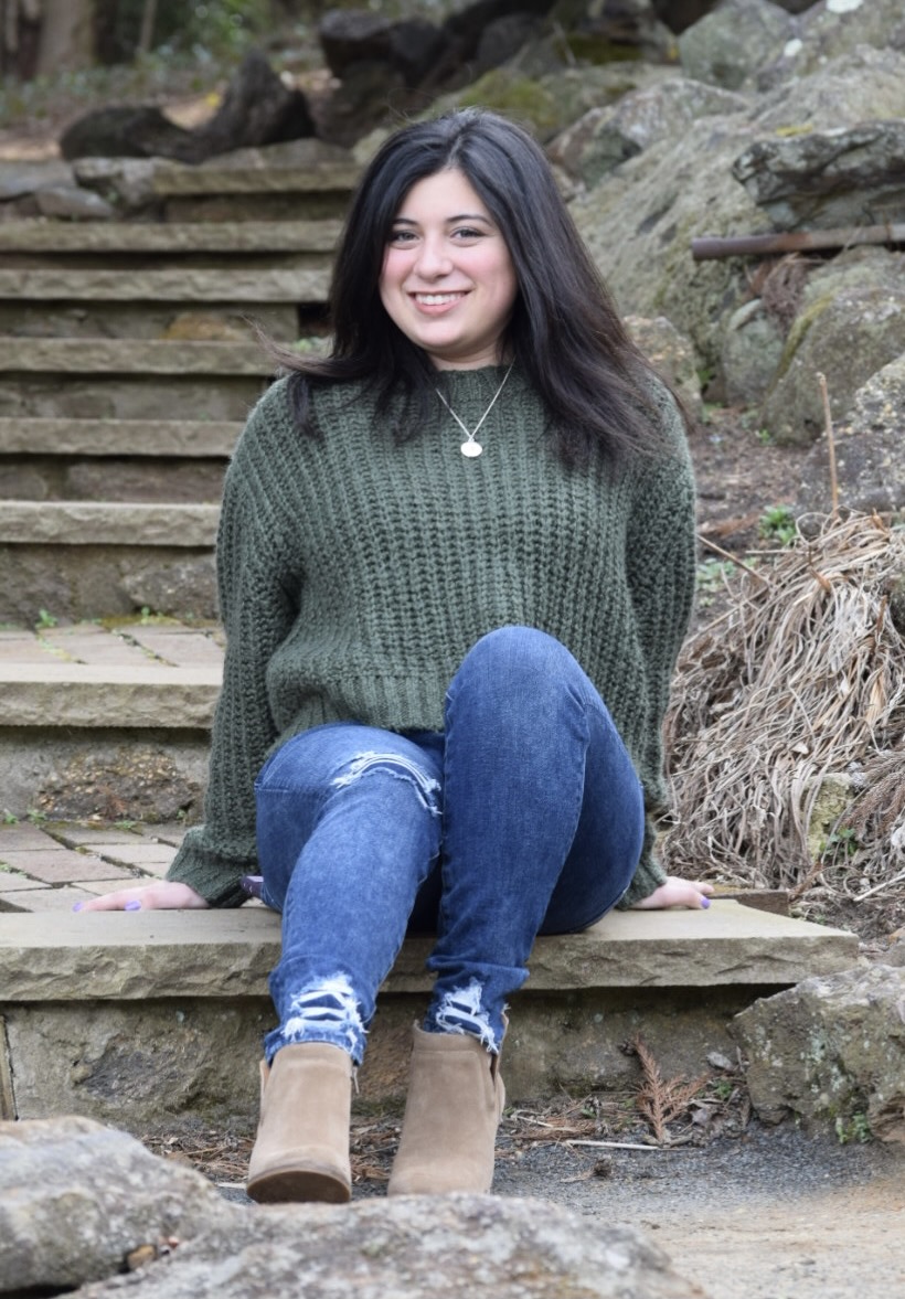 A light-skinned woman with medium length dark hair sits at the base of an outdoor, stone stairway.