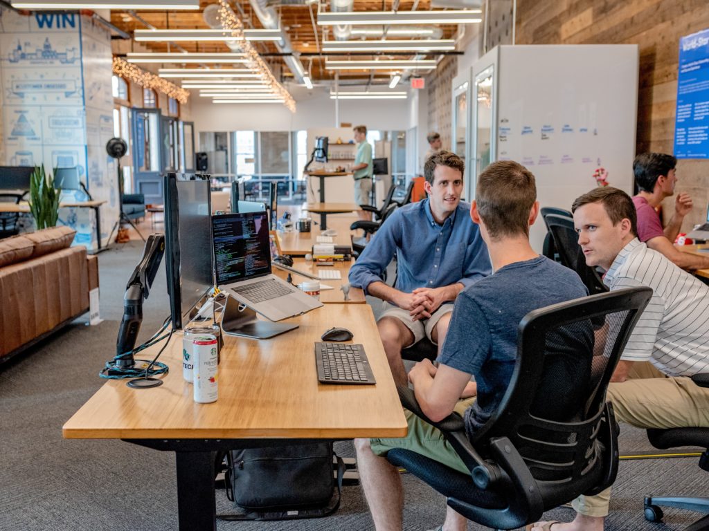 Three people sitting and talking in front of an adjustable desk with a laptop, external monitor, and keyboard.