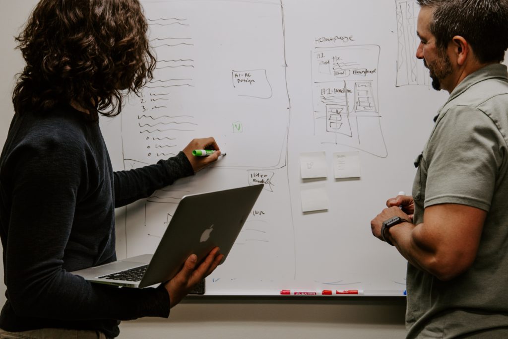 Two people stand at a white board. One is holding an open laptop while writing on the board with a green marker.