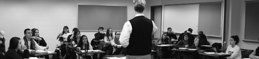 A light-skinned man is shown from the back and is standing at the front of a classroom. He looks out upon many young people of varying gender and skin tone who are seated in individual desks. All of the young people are looking up and toward the front of the classroom. 