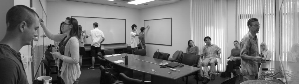 From the left side of the image: a light-skinned man is holding a coffee cup and looking downward. A light-skinned young person is facing and writing on a whiteboard on the left wall. Another light-skinned young person is standing to the first person's right and watching the writing. In the back of the room, two light-skinned young people stand next to each other and write on another whiteboard. To their right two medium-skinned young people stand facing a whiteboard on the right wall of the room. One of the people is writing on the whiteboard while the other observes from behind, his right hand rubbing his head. To their right: three light-skinned people and a medium-skinned person are seated in front of a window and observe a light-skinned man as he faces and writes on a whiteboard placed on the wall at the front of the room. 