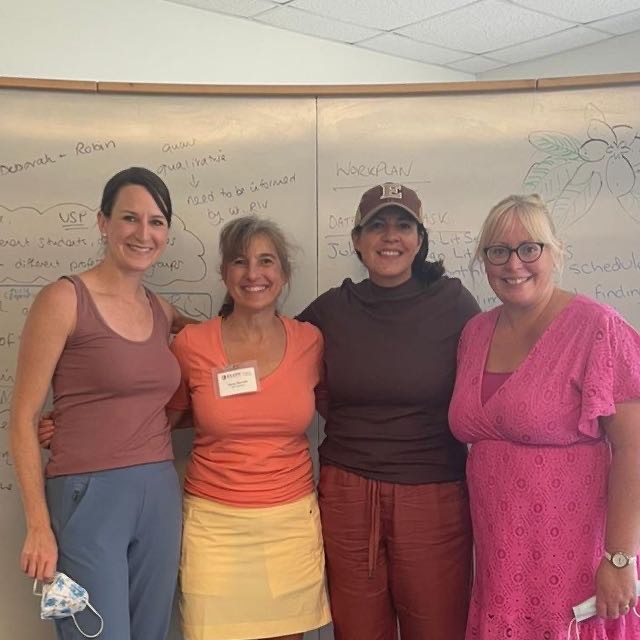 Four women stand arm-in-arm in front of a whiteboard filled with writing. They are smiling at the camera.