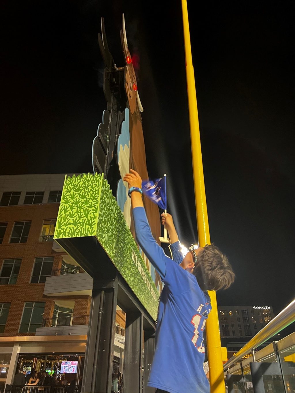 A photo of a boy with glasses wearing a blue jersey reaching up with flag toward a large billboard of a bull with eyes lit up and smoke coming from nose.