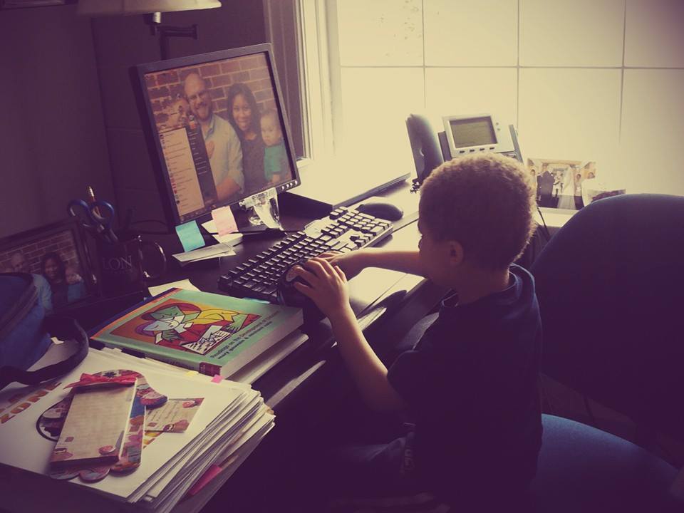 A young black boy sits at a desk crowded with books, papers and photographs. He has his left hand on a ball and his right on the keyboard.