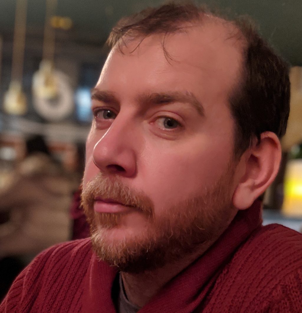 Headshot of a light-skinned male, Chris Trimby. He has a beard and mustache and wears a red sweater.