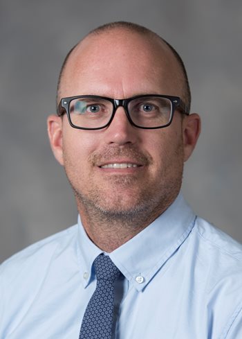 White man with glasses, wearing a blue shirt and navy blue tie.