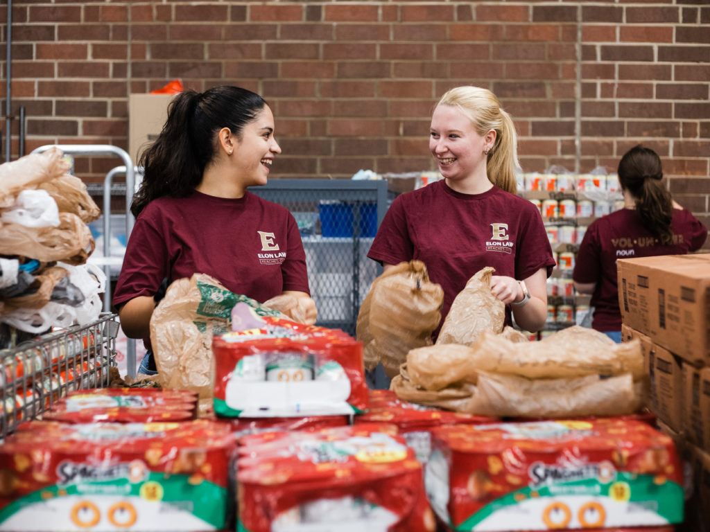 Two students look at each other, laughing. They both wear Elon Law T-shirts, and they are holding bags and standing near a table filled with packages of Spaghettio’s.