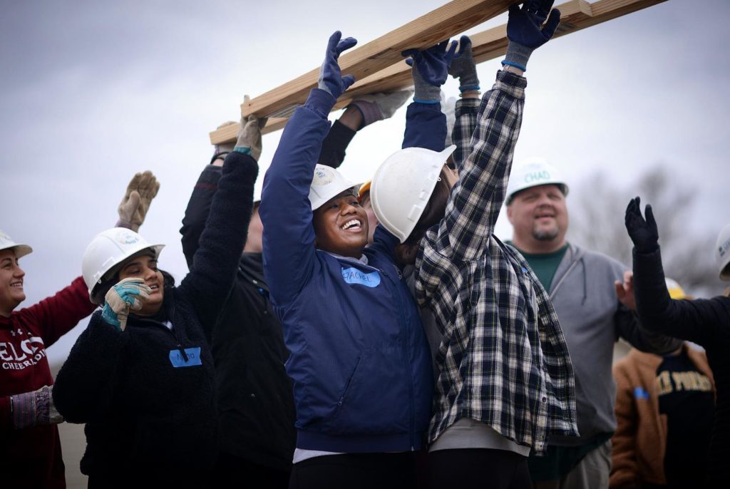 Several people wearing hard hats and heavy gloves lift pieces of lumber above their heads. Several are smiling or laughing.