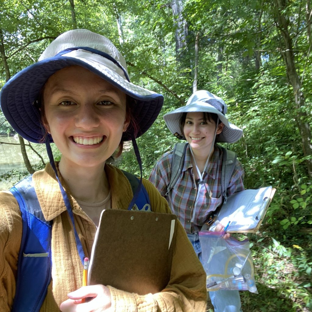 Two students stand in a forest, smiling toward the camera. They wear wide-brimmed hats and backpacks, and both are carrying clipboards and pencils. One student carries a ziplock bag filled with small objects.