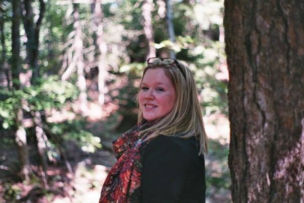 Woman standing in a wooded area. She's wearing a black jacket and a scarf with various shades of red. She has glasses on top of her head, and she's looking back over her shoulder.