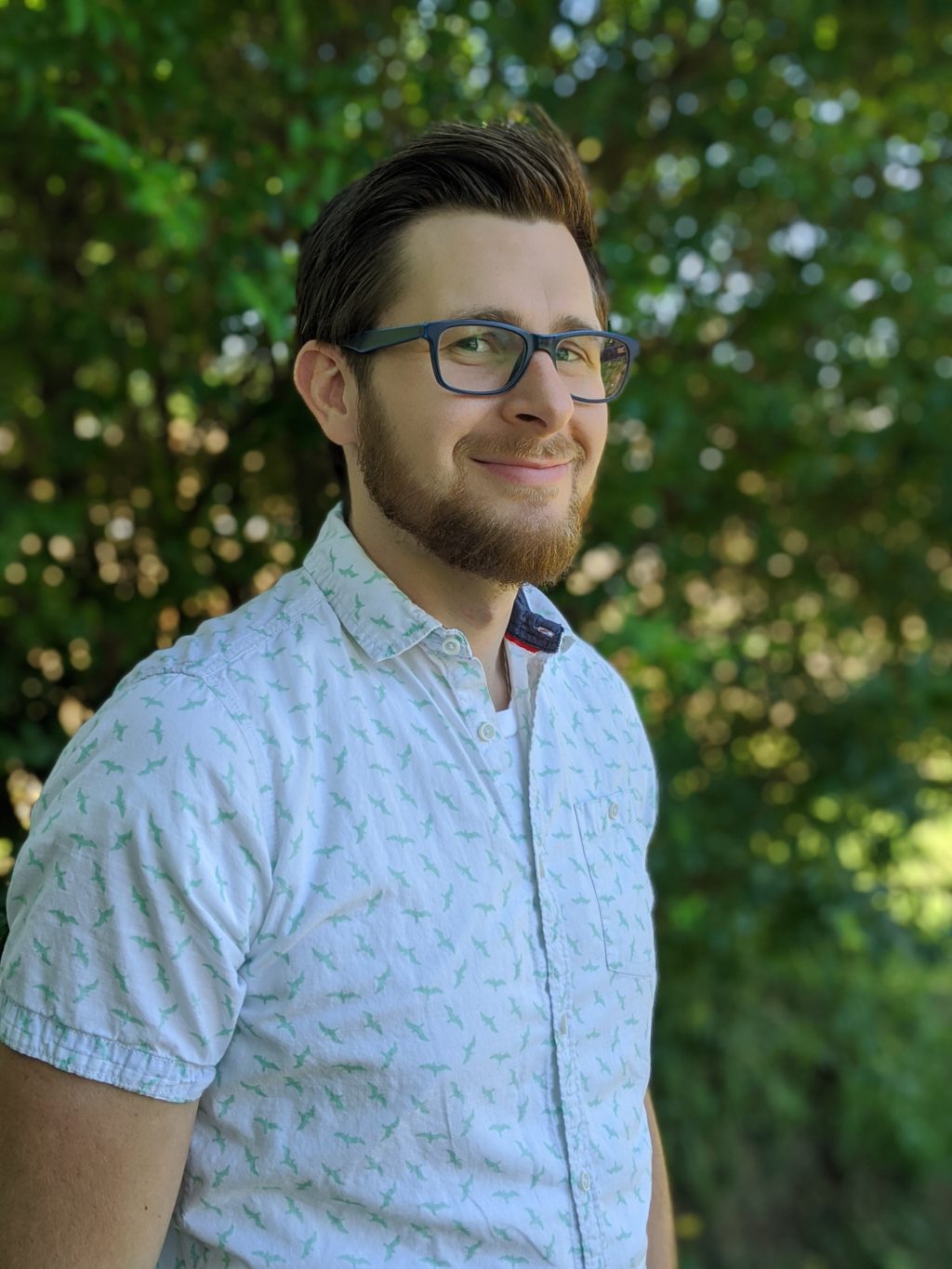 A light-skinned man with a beard and glasses stands in front of greenery.