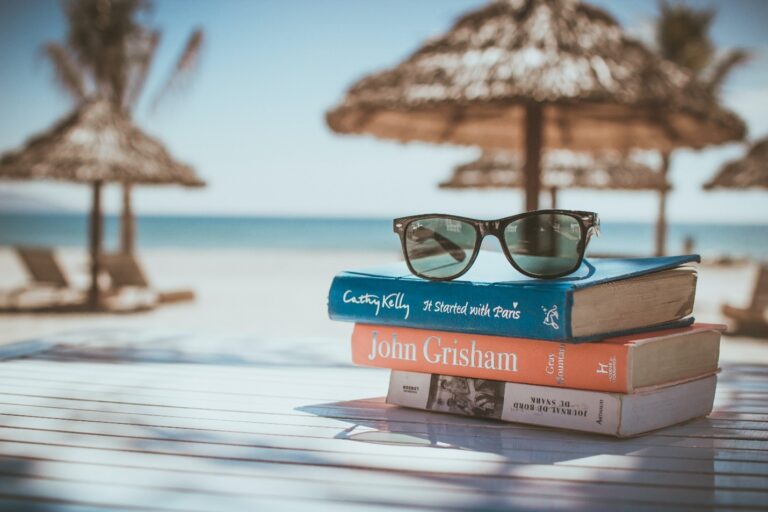 A pair of sunglasses on top of a three stacked books on a table with a beach in the background.