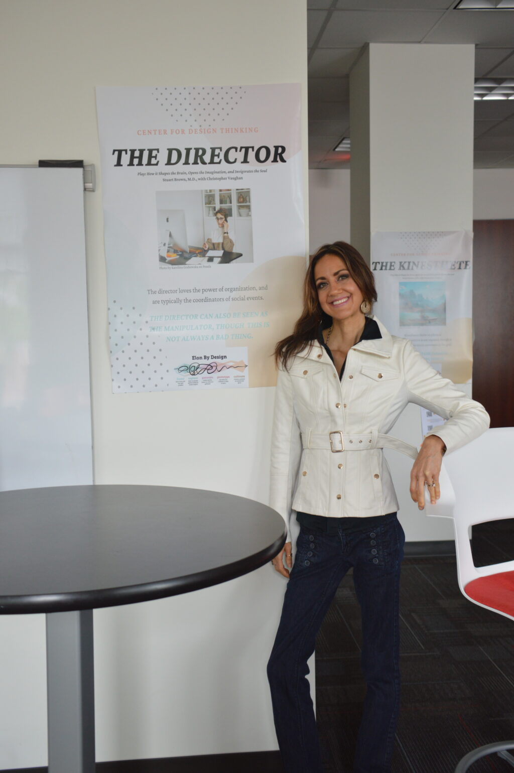 A woman stands by a high top table with her arm resting on a nearby bar-height chair. On the wall behind her, a poster reads, "The Director."