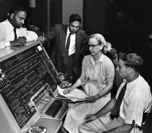 A black and white photo of a woman wearing a dress, cardigan, and dark glasses, sitting at a large computer terminal. Three men stand and sit around her. All are looking toward the computer.