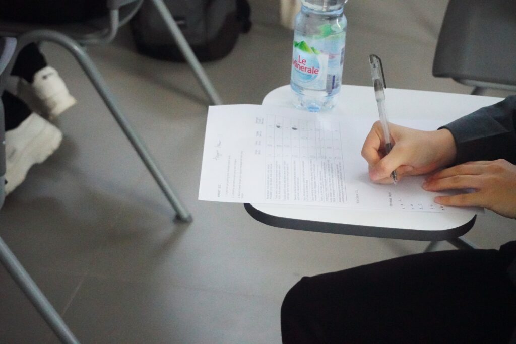 The hands of a person are visible writing on a paper at a classroom desk. A water bottle also sits on the desk. Another row of students is visible on the left side of the photo.