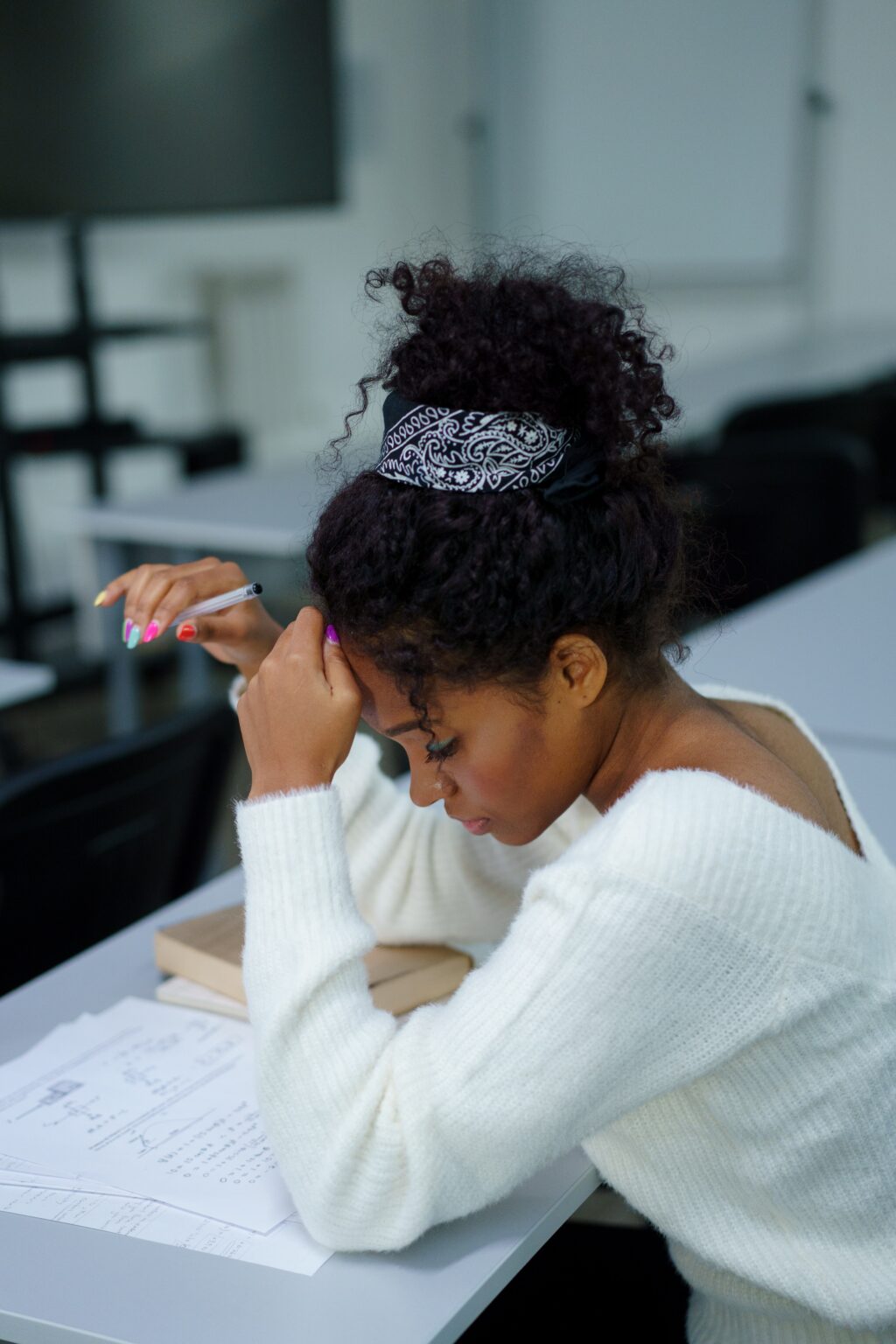 A student sits at a desk. She looks down at a few papers on the desk, which are filled with writing. She has one hand up to her forehead, and the other holds a pencil.