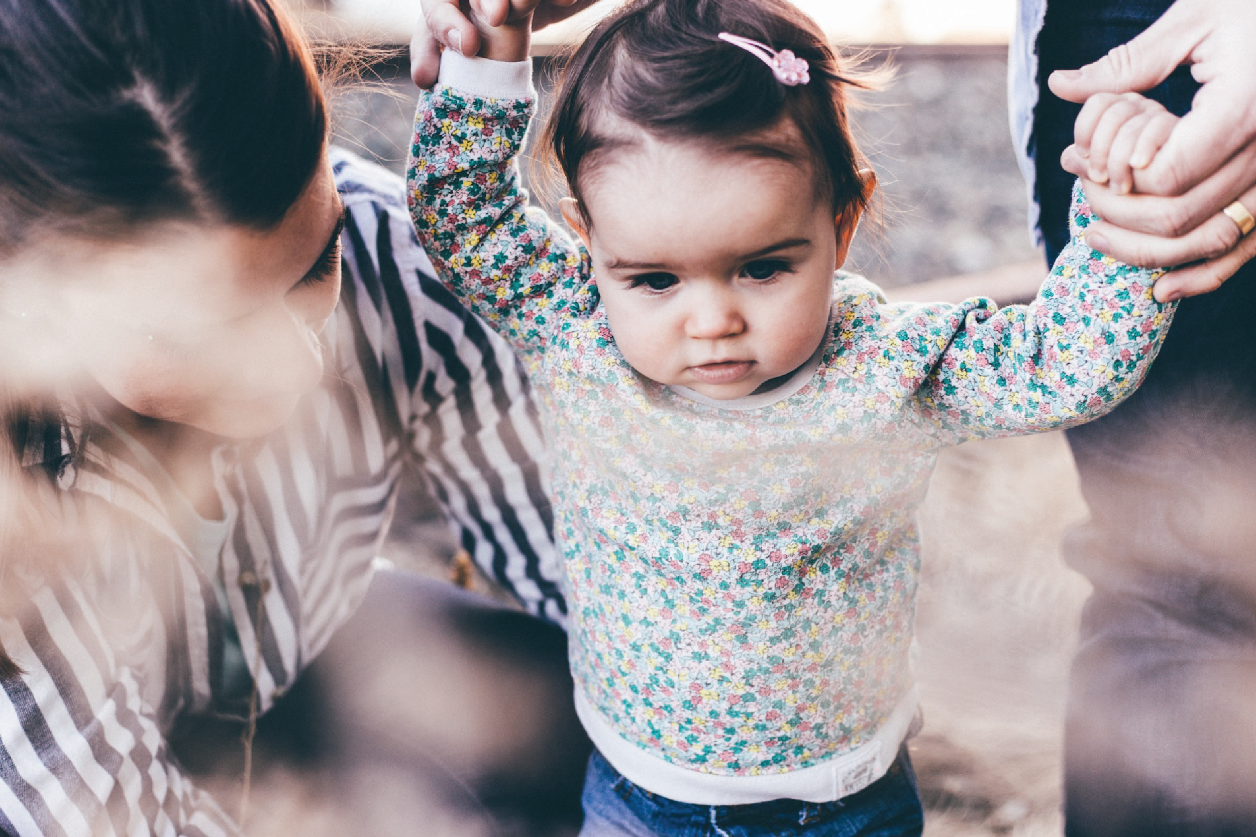 A baby in a flowered dress walks, with a parent holding their hands. A woman is visible next to the child.