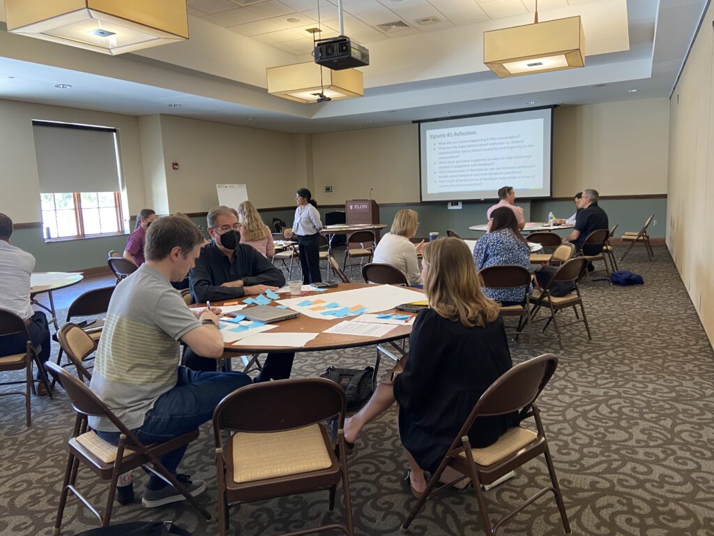 People sit at round tables, talking. Each table has several big sheets of paper covered with different colored, square sticky notes.