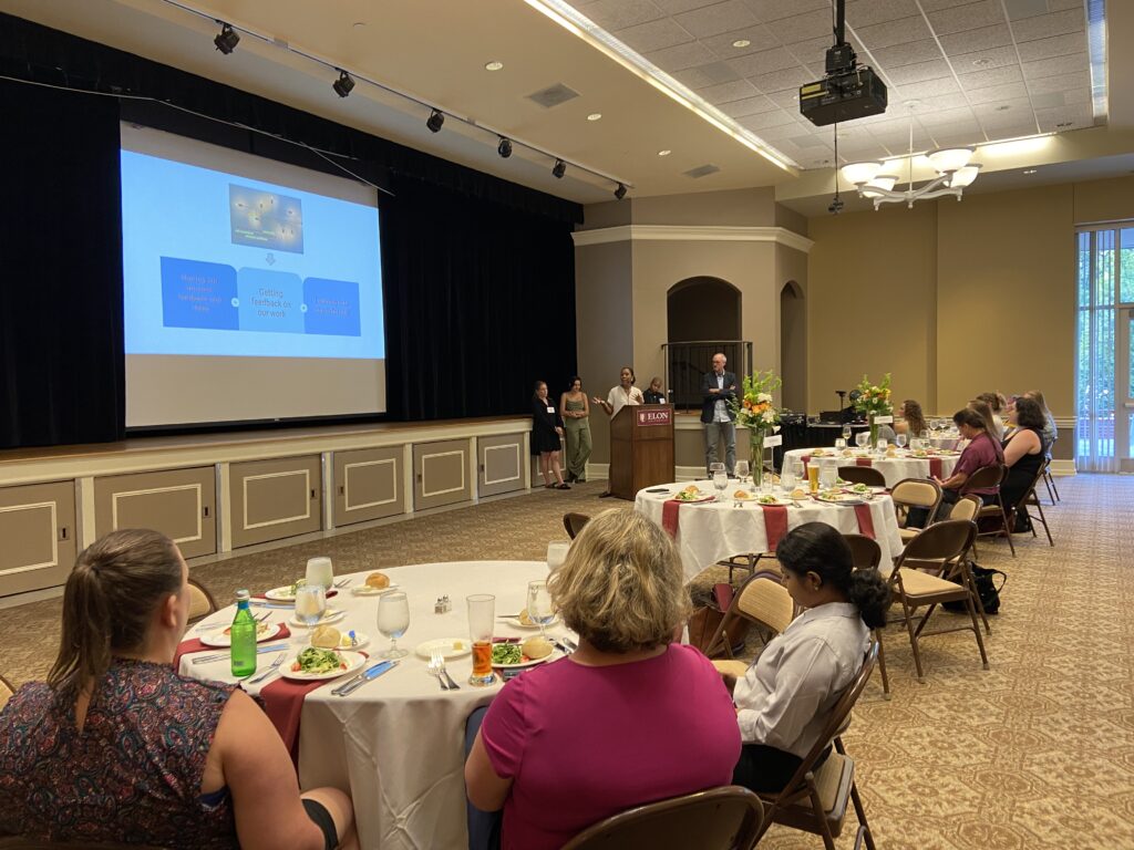 Five people stand near a podium in the distance. One gestures towards a presentation slide on a screen. People at four round tables look at the presenters. Some are taking notes.