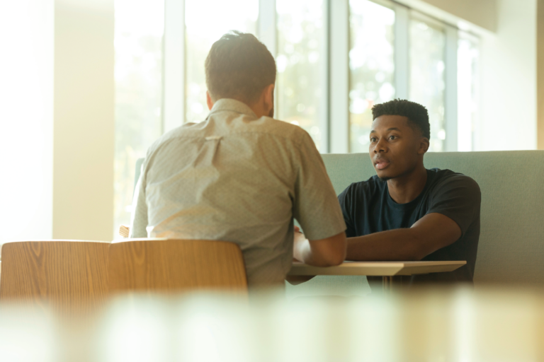 A young, dark-skinned man sits at a table across from a light-skinned man.