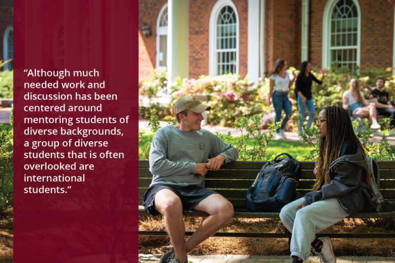 Two students sit on a bench talking. An academic building and more students are visible in the background. Text overlay reads, "Although much needed work and discussion has been centered around mentoring students of diverse backgrounds, a group of diverse students that is often overlooked are international students."