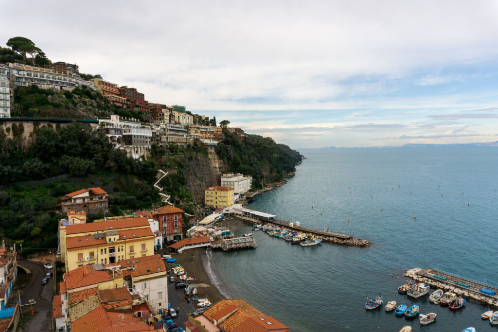 A small bay with a marina filled with boats is surrounded by buildings along a tall rocky shoreline.
