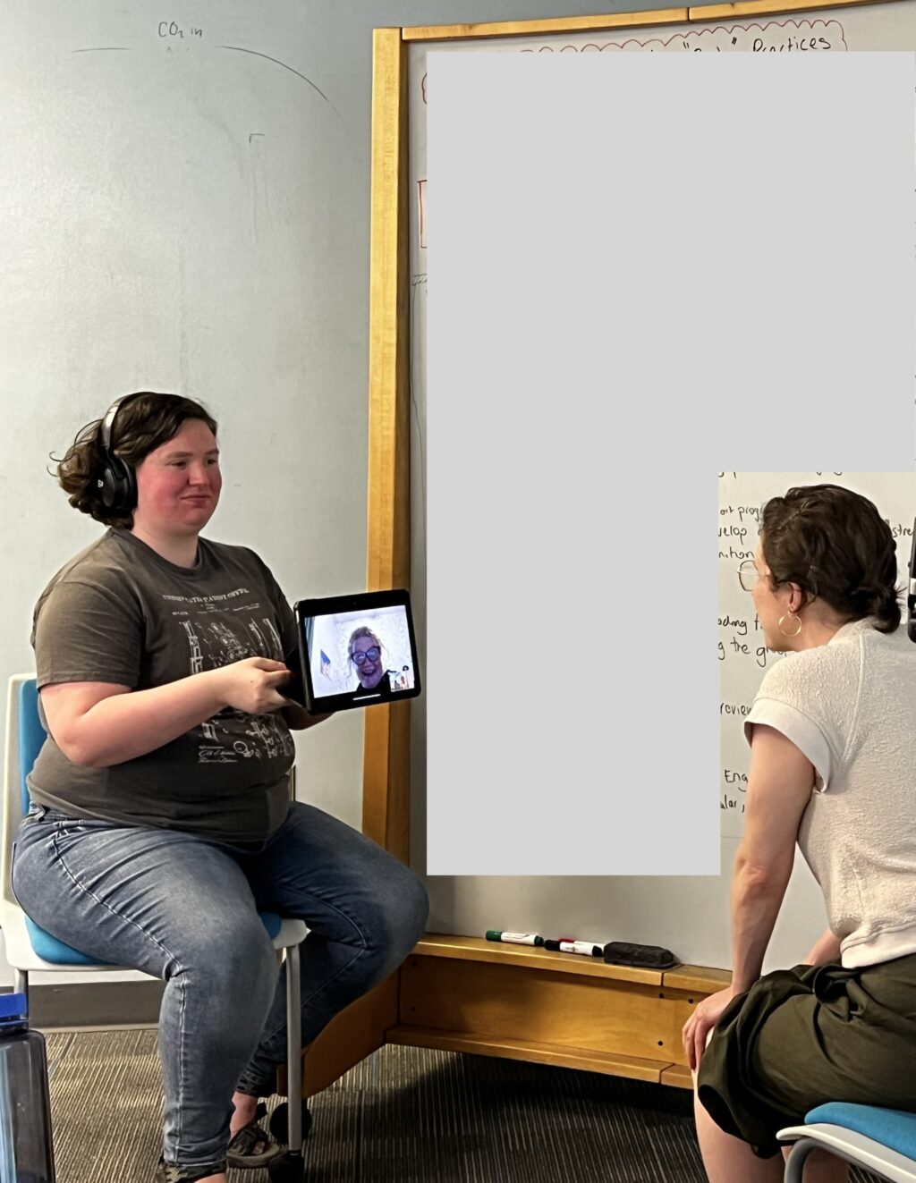 Photo of Kaz Kelly sitting in chair, holding an iPad so that a person joining remotely via the iPad can visit with a person sitting across from Kaz
