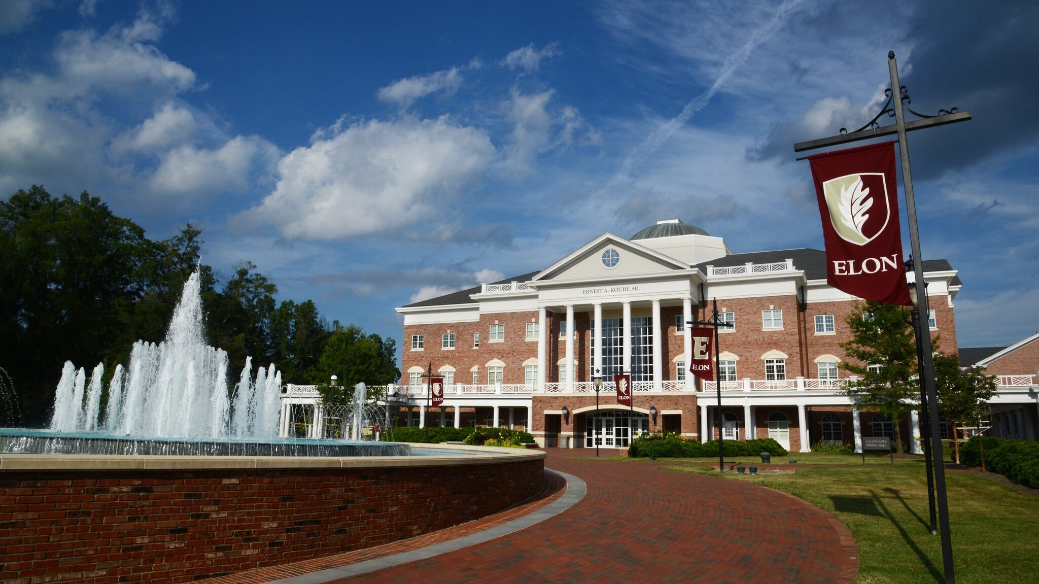 Elon University. Two story brick building with white pillars and a fountain in front with blue sky.