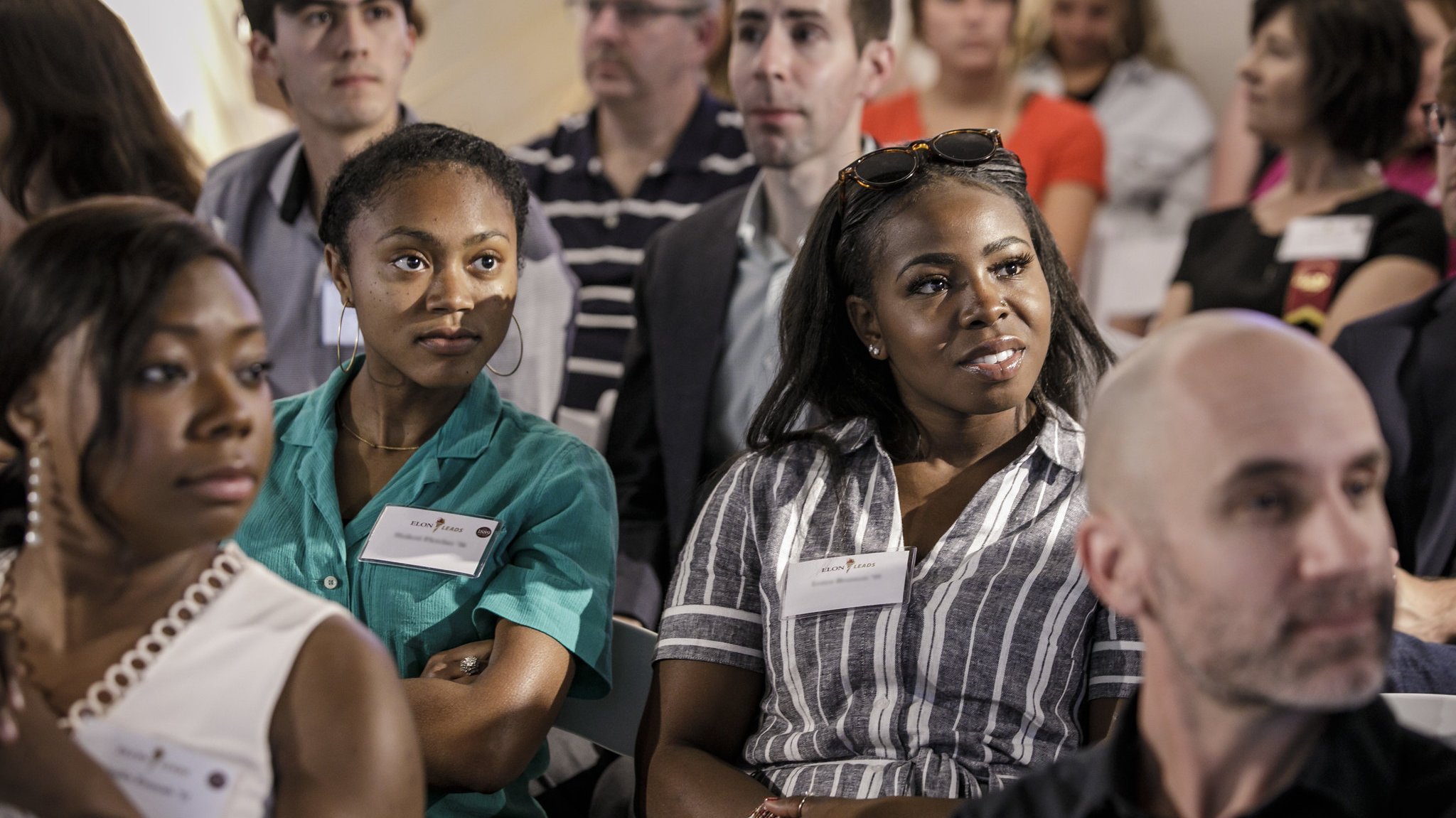 Two Black women students look beyond the camera. People sitting in a conference style audience surround them.