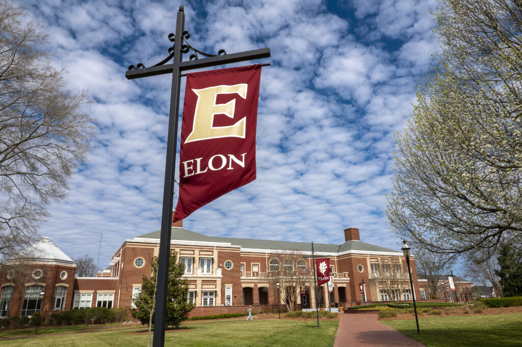 In the foreground, an Elon banner hangs on a decorative metal pole. Moseley Center, a large brick building, is in the background.