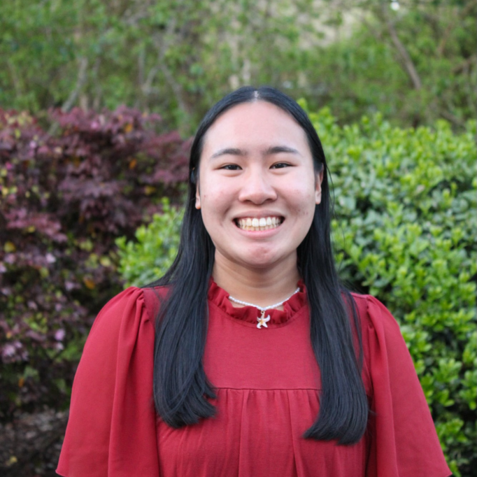 A picture of a young woman in a red shirt smiling in front of green bushes.