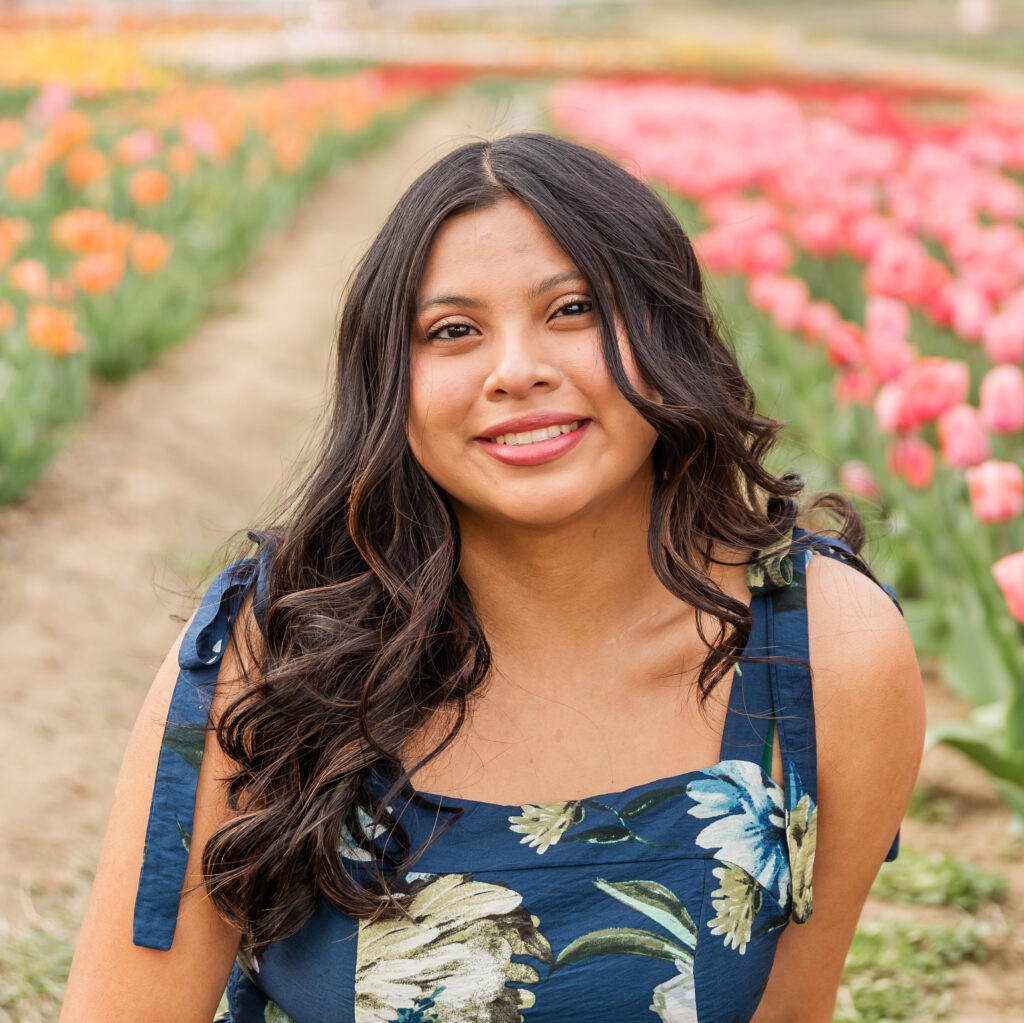 A young woman with long dark brown hair in a navy floral dress smiling with pink and orange flowers in the background