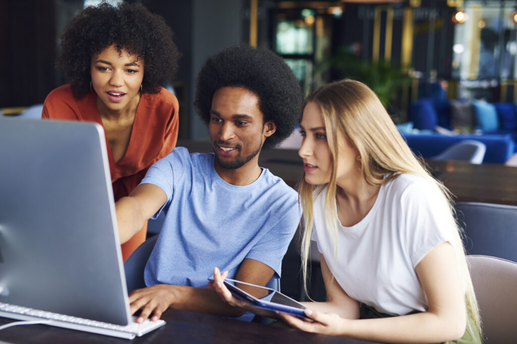 Three young coworkers gather in front of a computer. One holds a tablet.