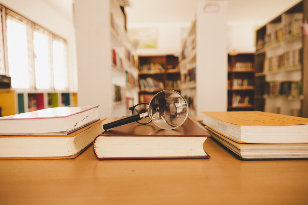 Three piles of books on a table, with library stacks in the background. On the middle pile of books are a magnifying glass and a pair of glasses.