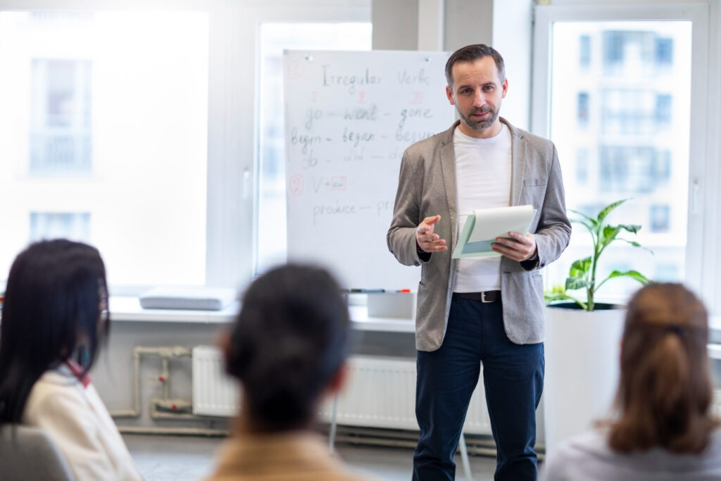 Man stands holding a notepad and talking to three people who are seated facing him. Behind him are an easel with a large note pad, windows, and a plant.